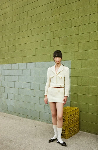 An adult female in a cream skirt suit poses outdoors against a two-tone green brick wall, next to stacked yellow crates.