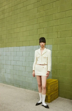 An adult female in a cream skirt suit poses outdoors against a two-tone green brick wall, next to stacked yellow crates.