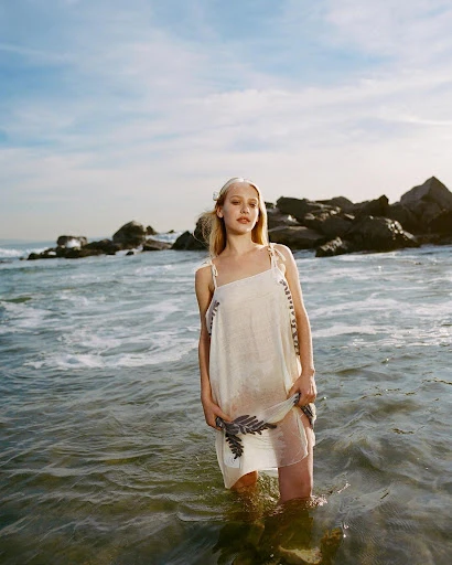 A woman stands in ocean water wearing a summer dress with a fern design, rocky coastline and blue sky with clouds in the background.