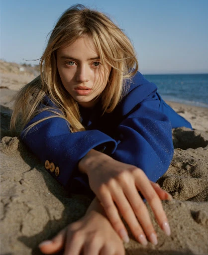 Eye-level shot of a blonde woman in a blue blazer lying on a sandy beach against a backdrop of clear blue sky and ocean.