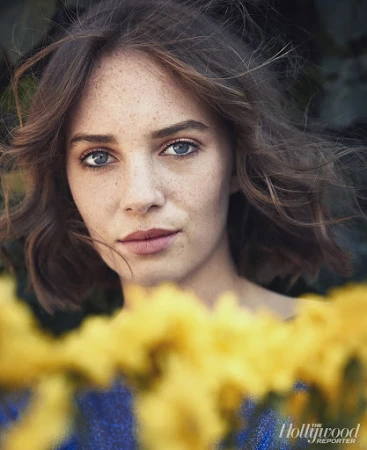 Close-up portrait of a woman with freckles and brown hair, framed behind out-of-focus yellow flowers against dark foliage.