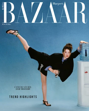 Ever Anderson holding a glass of water and kicking high with one leg while leaning against a water cooler in a blue studio setting.