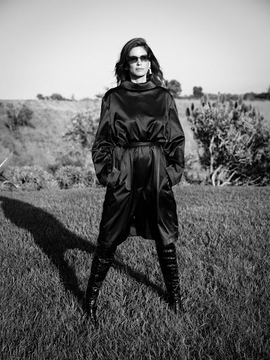 A woman in a black coat and boots stands in a grassy field in a black-and-white fashion portrait, looking at the camera.