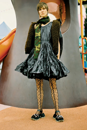 Model standing outdoors wearing a shiny dark tiered dress, patterned tights, and a fuzzy jacket draped over shoulders.