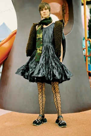Model standing outdoors wearing a shiny dark tiered dress, patterned tights, and a fuzzy jacket draped over shoulders.