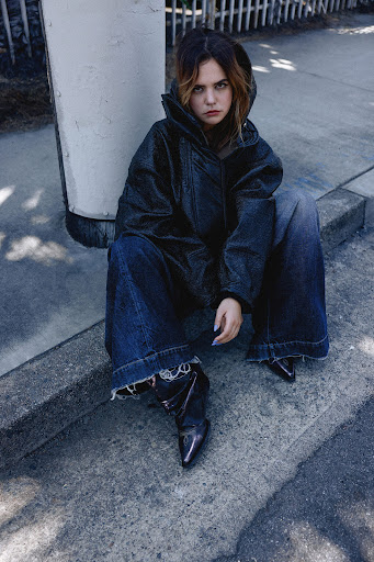 Fashionable woman in denim jacket and jeans sits on concrete outside with natural light, showcasing street style and modern apparel.