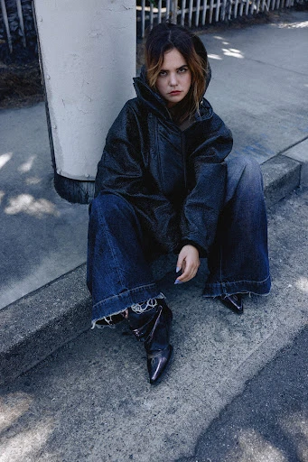 Fashionable woman in denim jacket and jeans sits on concrete outside with natural light, showcasing street style and modern apparel.