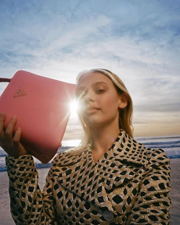 Blonde woman holding a pink handbag against a beach and sunset sky, wearing a patterned coat.