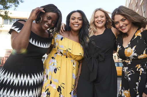 Group of four women standing close together outdoors, smiling at the camera, wearing casual dresses and fashionable outfits in sunlight.
