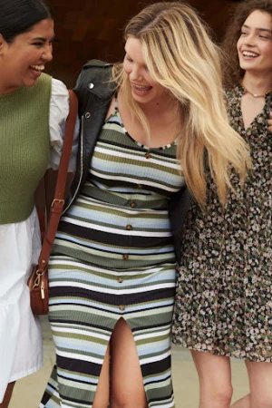 Three women of mixed ethnicities smile outdoors. The woman in the middle wears a black jacket and a vertically striped dress.