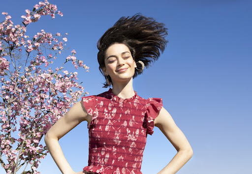 Adult woman smiling outdoors near flowering tree. The model is wearing a red printed dress with a blue sky in the background.