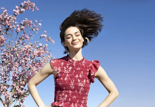 Adult woman smiling outdoors near flowering tree. The model is wearing a red printed dress with a blue sky in the background.