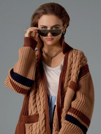 Woman wearing wraparound sunglasses and chunky cable-knit cardigan against a solid grey background