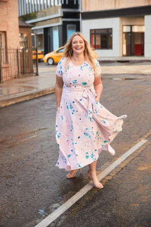 Smiling blonde woman in a pink floral midi dress walking outdoors on a city street with a yellow taxi in the background.