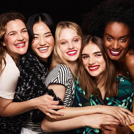 A group of five smiling women of varying ethnicities are closely grouped together in a studio portrait against a dark backdrop.