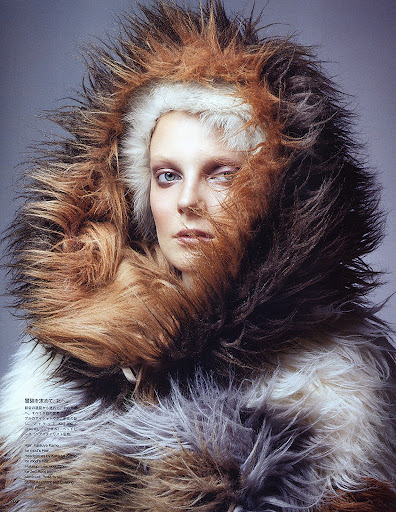 Close-up of a Caucasian female model with fair skin and light eyes, wearing a large, multi-toned brown and white fur coat with a hood.