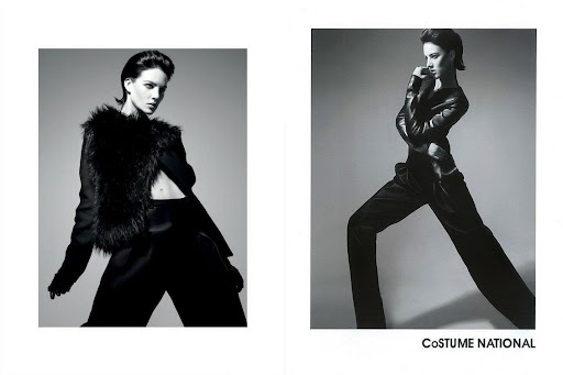 Black and white fashion shot of a model wearing a fur vest and black suit, posing against a grey backdrop with dramatic lighting.