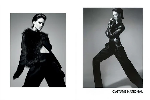 Black and white fashion shot of a model wearing a fur vest and black suit, posing against a grey backdrop with dramatic lighting.