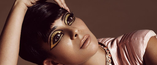 A woman with artistic gold eye makeup looks up with a neutral expression, wearing a light pink top and beaded necklace in a studio shot.