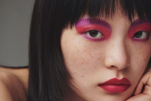 Asian woman with bold pink and red eyeshadow and lipstick, black hair, and freckles poses in front of a grey background.
