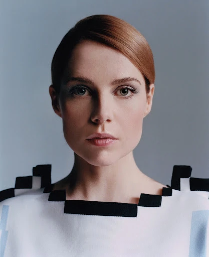 Portrait of an adult female with auburn hair and light eyes wearing a white top with black square embellishments against a light gray background.