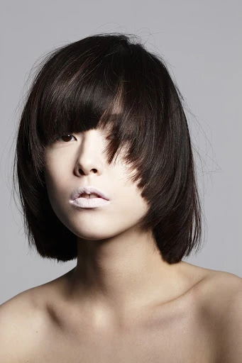 Headshot of a young adult woman with dark, short hair, light skin, and pale pink lipstick, posing in a photo studio with a grey background.