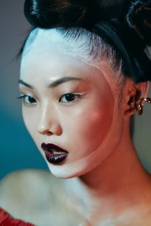 Close-up portrait of an East Asian woman with avant-garde white face paint, dark glossy lips, and elaborate dark updo styled by Lisa Houghton.