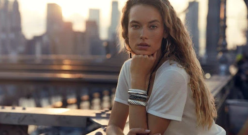 Portrait of a young woman in a white t-shirt wearing bracelets, posing in front of a city skyline with a golden sunset.