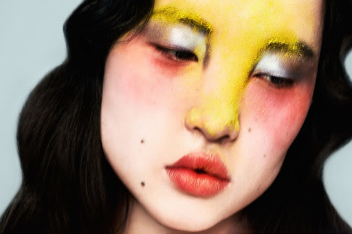 Close-up portrait of an Asian woman with bold yellow and silver makeup, red cheeks, and dark wavy hair against a plain background.