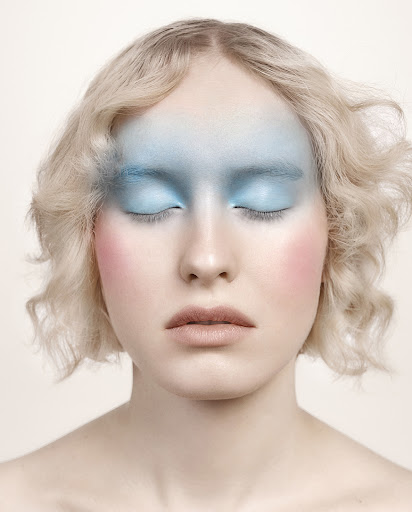 Close-up studio shot of a woman with fair skin, closed eyes, and wavy blonde hair, wearing pastel blue and pink makeup.