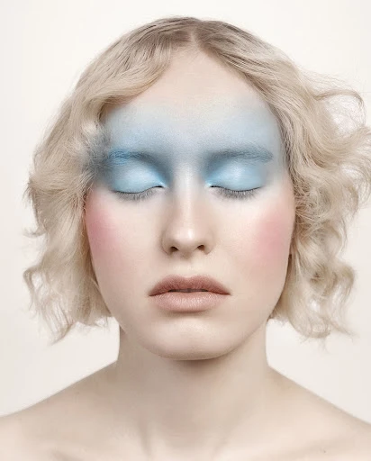 Close-up studio shot of a woman with fair skin, closed eyes, and wavy blonde hair, wearing pastel blue and pink makeup.