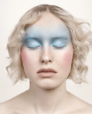 Close-up studio shot of a woman with fair skin, closed eyes, and wavy blonde hair, wearing pastel blue and pink makeup.