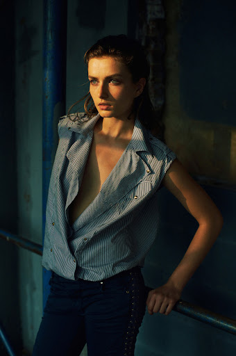 Portrait of a young adult woman with an open striped vest leaning on a metal rail, blue wall in the background, bathed in golden light.