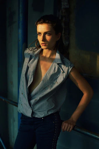Portrait of a young adult woman with an open striped vest leaning on a metal rail, blue wall in the background, bathed in golden light.