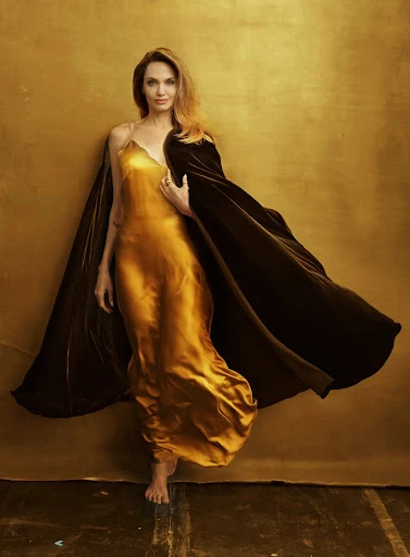 Angelina Jolie poses barefoot in a golden dress with a brown cape against a gold backdrop, capturing a moment of elegance and grace.