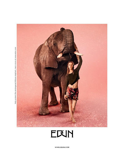 A model poses next to a large elephant in a studio setting with a pink background, promoting wildlife conservation.