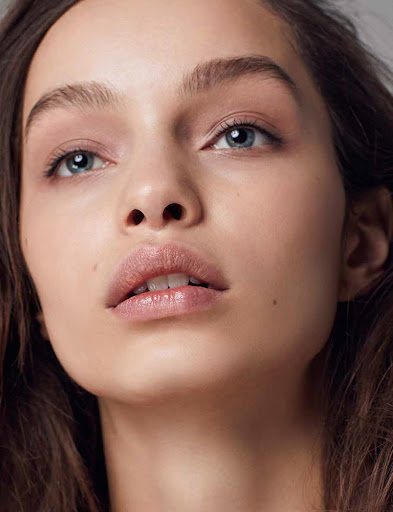 Close up beauty portrait of a young woman with natural makeup, light blue eyes, and glossy lips against a neutral background