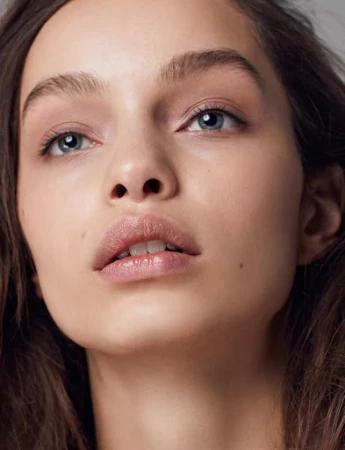 Close up beauty portrait of a young woman with natural makeup, light blue eyes, and glossy lips against a neutral background