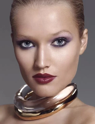 Close-up portrait of a woman with blonde hair, red lipstick, and dramatic eye makeup, wearing a large gold and silver choker necklace.