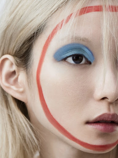 Close-up portrait of a fair-skinned woman with blonde hair, striking blue eyeshadow, and a bold red painted circle around her face.