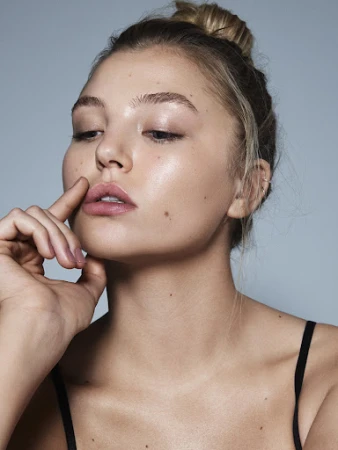 Close-up beauty portrait of a woman with dewy skin and natural makeup touching her lip against a soft grey background.