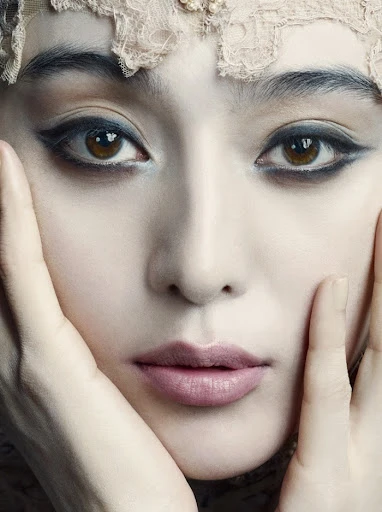 Close-up portrait of an adult woman with fair skin, dramatic eyeliner and light pink lipstick, with hands near her face.