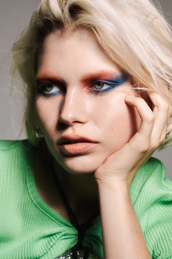 Close-up portrait of a young blonde woman with colorful eyeshadow and green top, looking serene with her hand resting on her chin.
