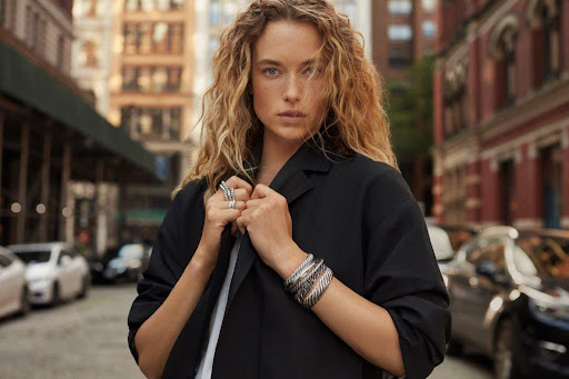 A fashionable woman with curly blonde hair wears jewelry and a black blazer on a city street with cars and brick buildings.