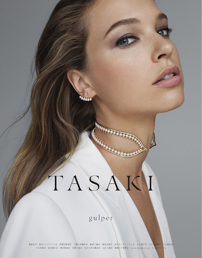Close-up of a woman wearing a white blazer and pearl jewelry, including a choker and earrings, against a gray background.