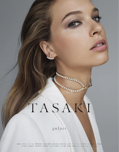Close-up of a woman wearing a white blazer and pearl jewelry, including a choker and earrings, against a gray background.