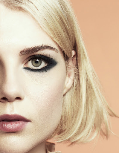 Close-up portrait of a young adult female with blonde hair and dark eye makeup against a peach background in a beauty editorial shot.