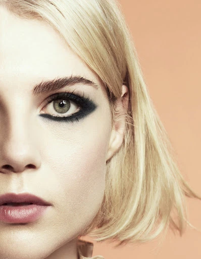 Close-up portrait of a young adult female with blonde hair and dark eye makeup against a peach background in a beauty editorial shot.