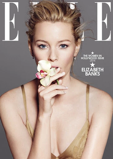 Elizabeth Banks poses on the cover of Elle Magazine with blonde hair and a flower held near her mouth against a grey background.
