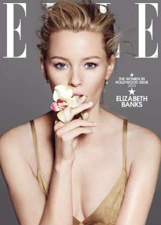 Elizabeth Banks poses on the cover of Elle Magazine with blonde hair and a flower held near her mouth against a grey background.